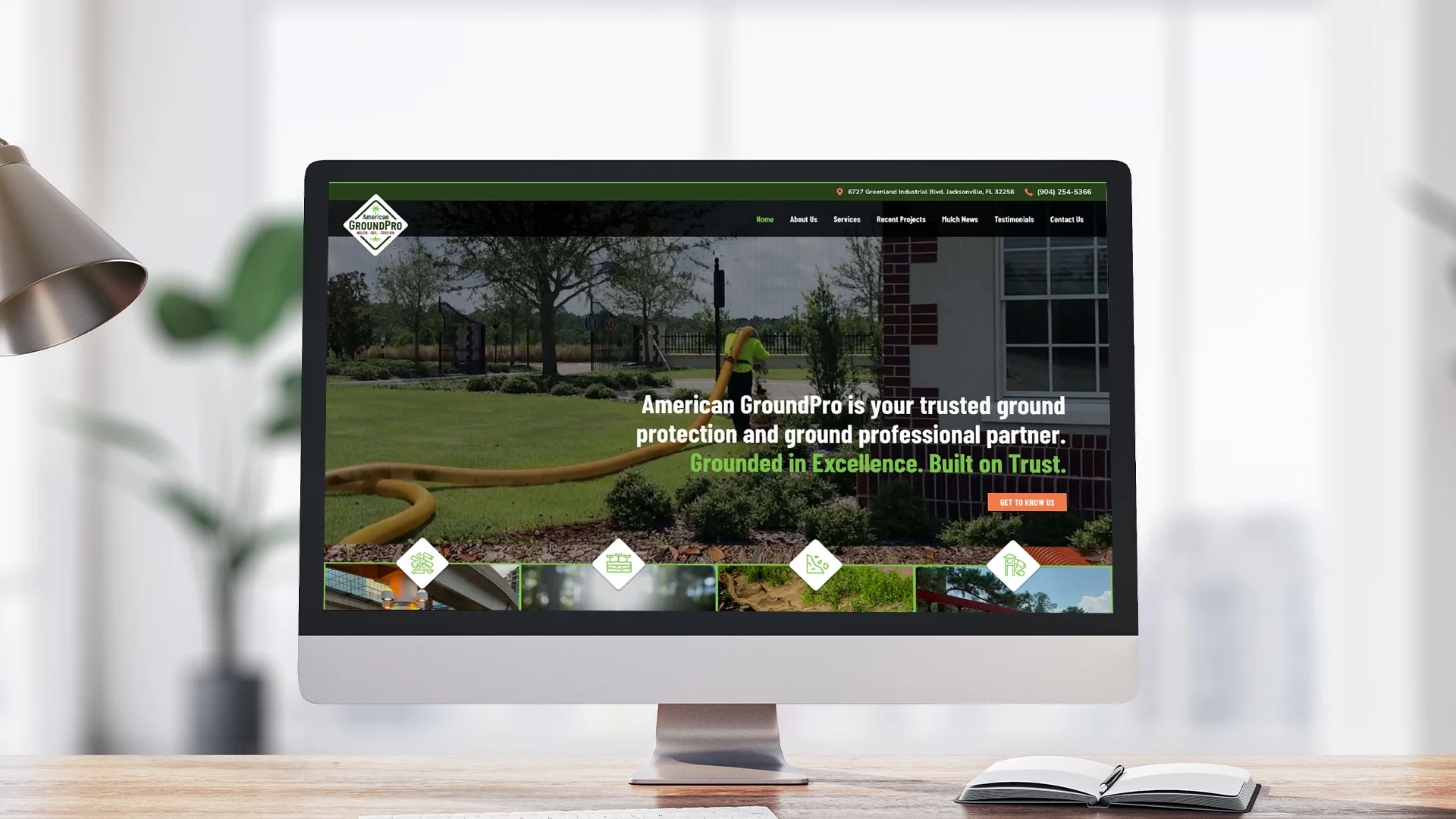 A computer monitor on a desk displays the American GroundPro website, featuring a lawn care service with the slogan Grounded in Excellence. Built on Trust. visible on the homepage.